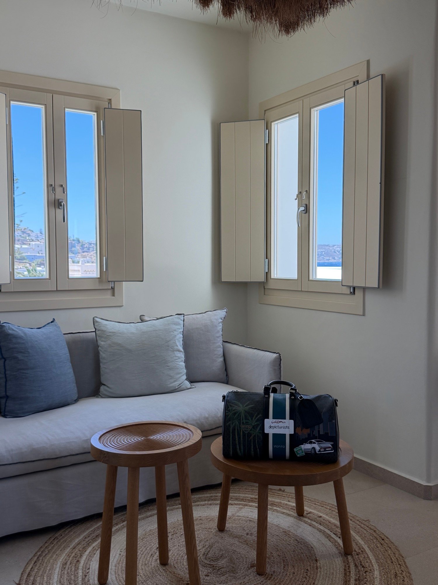 A sunlit hotel room with open shutters, a linen sofa and a travel bag on a wooden side table