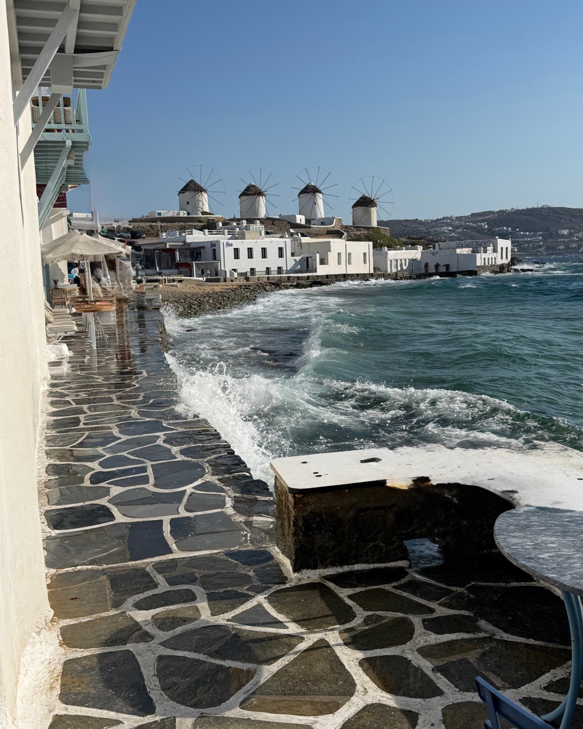 Waves reaching a stone terrace in Mykonos, with the windmills across the bay